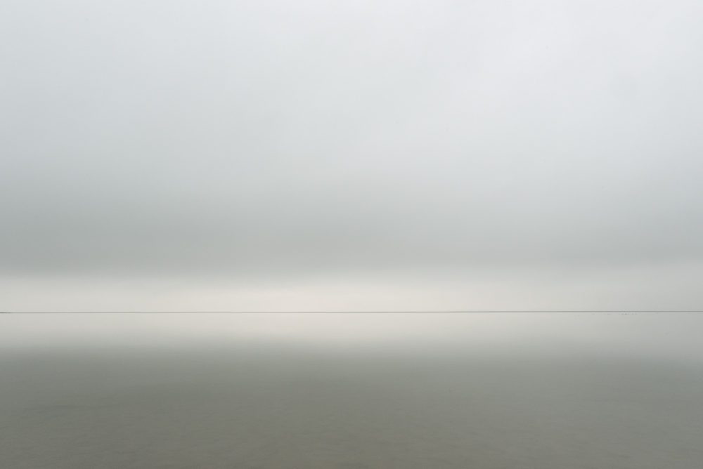A minimal photograph of a distant horizon looking across Morecambe Bay in the UK. The conditions are calm and cloudy.